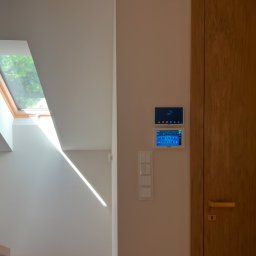 SmartMP Michał Polak - Dwa panele sterowania systemem inteligentnego domu zamontowane na ścianie obok drewnianych drzwi i włączników światła, z oknem dachowym w tle.