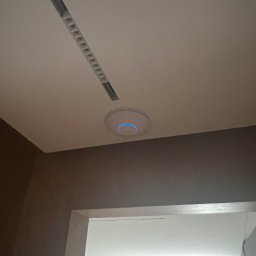 SmartMP Michał Polak - Zamontowany punkt dostępowy Wi-Fi z niebieską diodą na suficie w Radomiu, widoczny fragment listwy oświetleniowej i wejście do pokoju.