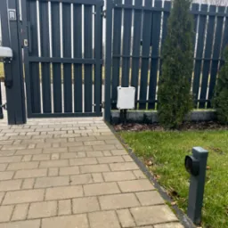 Nowoczesna, grafitowa brama posesji z automatyką, widoczny panel sterowania na słupku i skrzynka sterownicza przy trawniku, wyłożona kostką ścieżka.