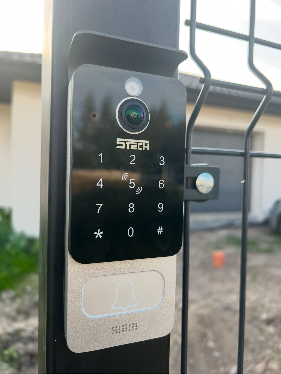 Nowoczesny wideodomofon z klawiaturą numeryczną i kamerą, zamontowany na czarnym słupku ogrodzenia panelowego, z widocznym logo producenta S-Tech.