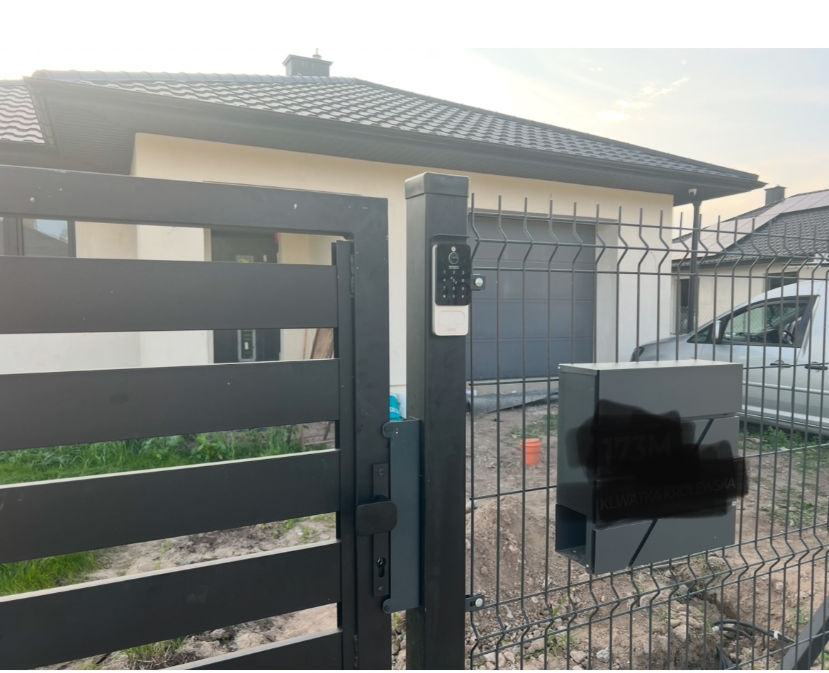 Nowoczesna, czarna brama posesji z panelem domofonu z klawiaturą numeryczną i skrzynką na listy, na tle domu z garażem i ogrodzeniem z siatki.