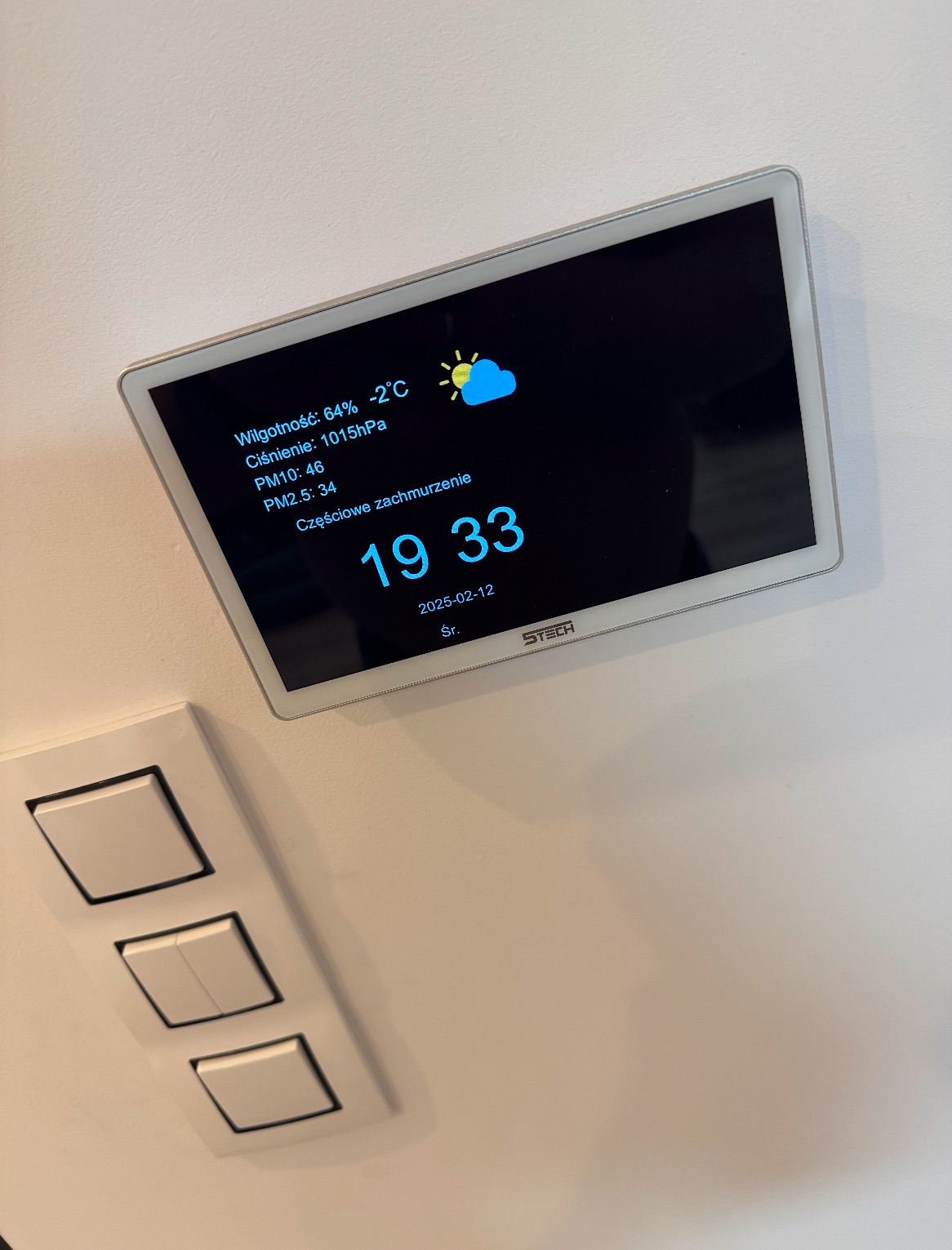 Panel sterowania inteligentnego domu na ścianie, wyświetlający dane o wilgotności, temperaturze, ciśnieniu, poziomie PM10 i PM2.5 oraz godzinę i datę, umieszczony obok włączników światła.