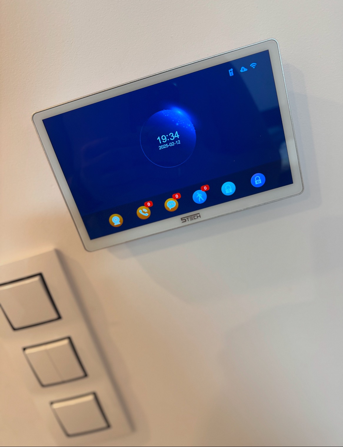 Panel sterowania inteligentnego domu zamontowany na ścianie, wyświetlający godzinę 19:34 i datę 2025-02-12, obok widoczne trzy włączniki światła.