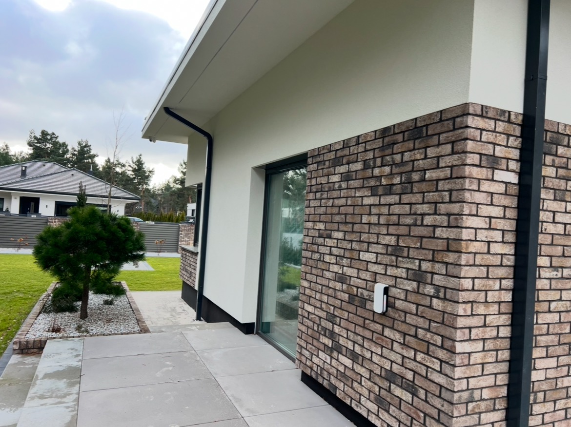 Nowoczesny dom jednorodzinny z elewacją z cegły klinkierowej w odcieniach brązu i beżu, z widocznym gniazdkiem na ścianie, szarą kostką brukową, czarną rynną i starannie utrzymanym trawnikiem...