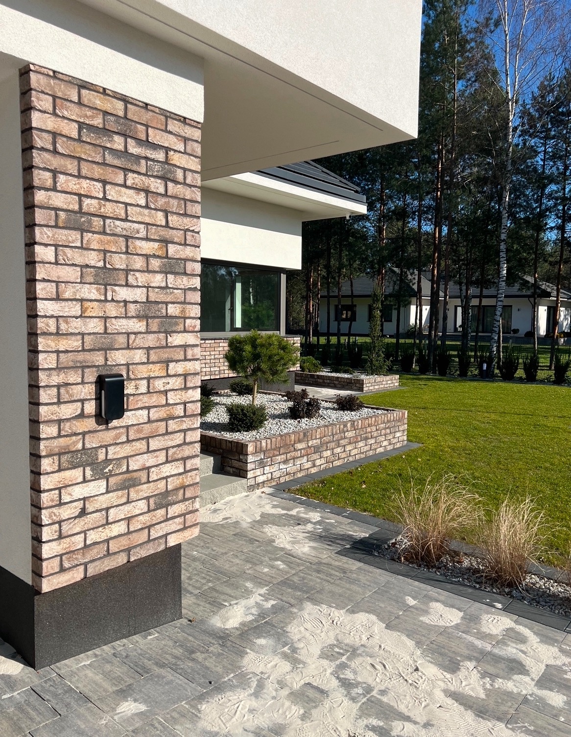 Nowoczesny dom jednorodzinny z elewacją z cegły klinkierowej, automatyką bramową i zadbanym trawnikiem, widok na skrzynkę na listy i fragment ścieżki z kostki brukowej.