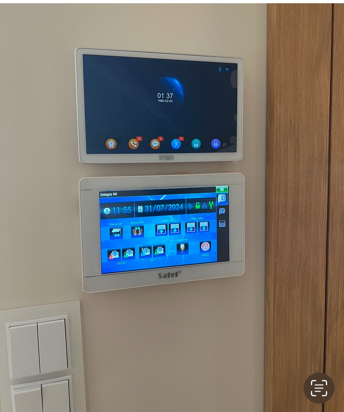 Dwa panele dotykowe inteligentnego domu zamontowane na ścianie obok włącznika światła i drewnianej framugi drzwi. Górny panel wyświetla godzinę i datę, a dolny panel marki Satel Integra 64 pokazuje...