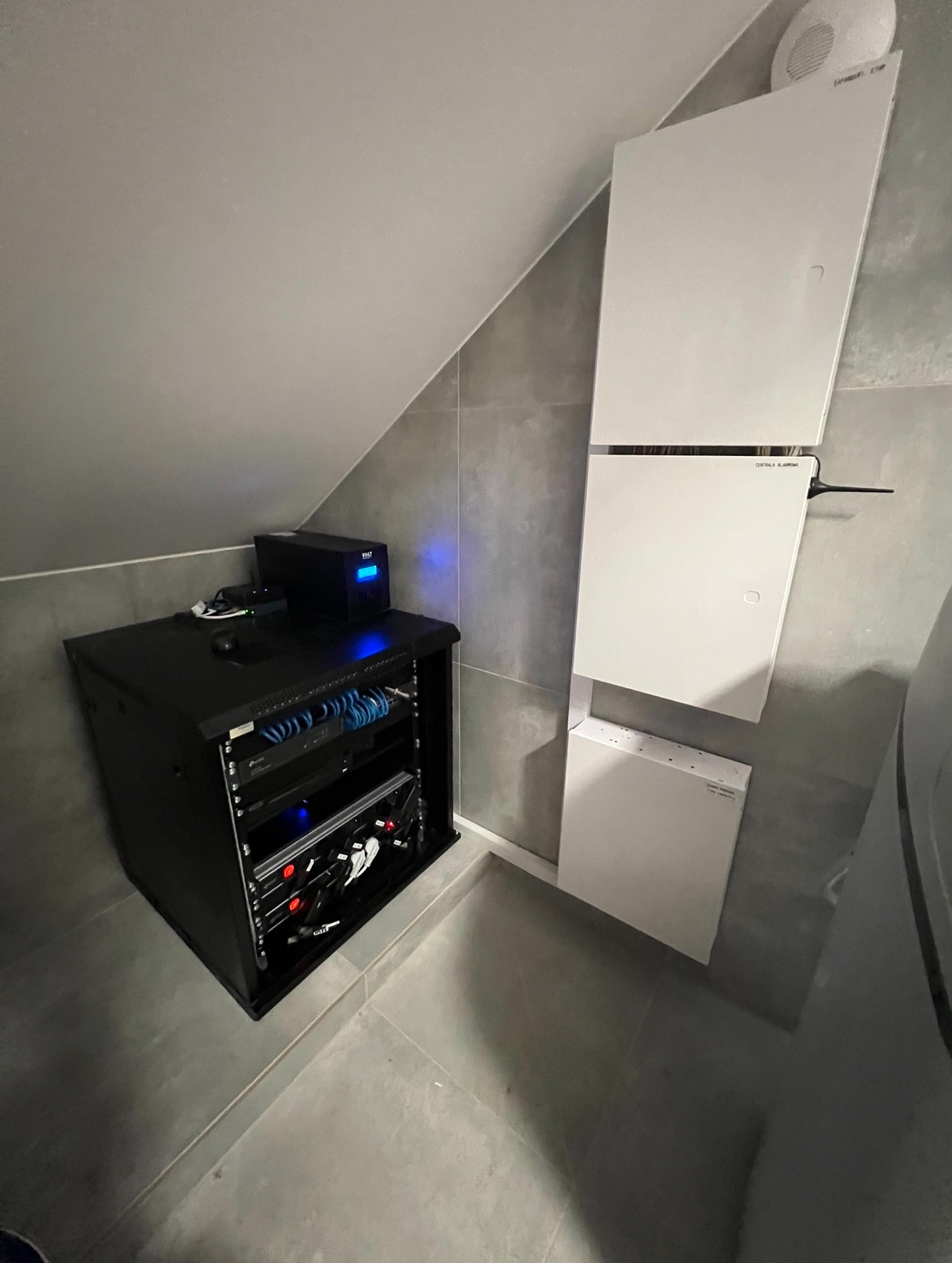 Czarna szafa rack z urządzeniami sieciowymi i zasilaczem awaryjnym UPS w rogu pomieszczenia z szarymi płytkami, obok białe skrzynki elektryczne na ścianie.