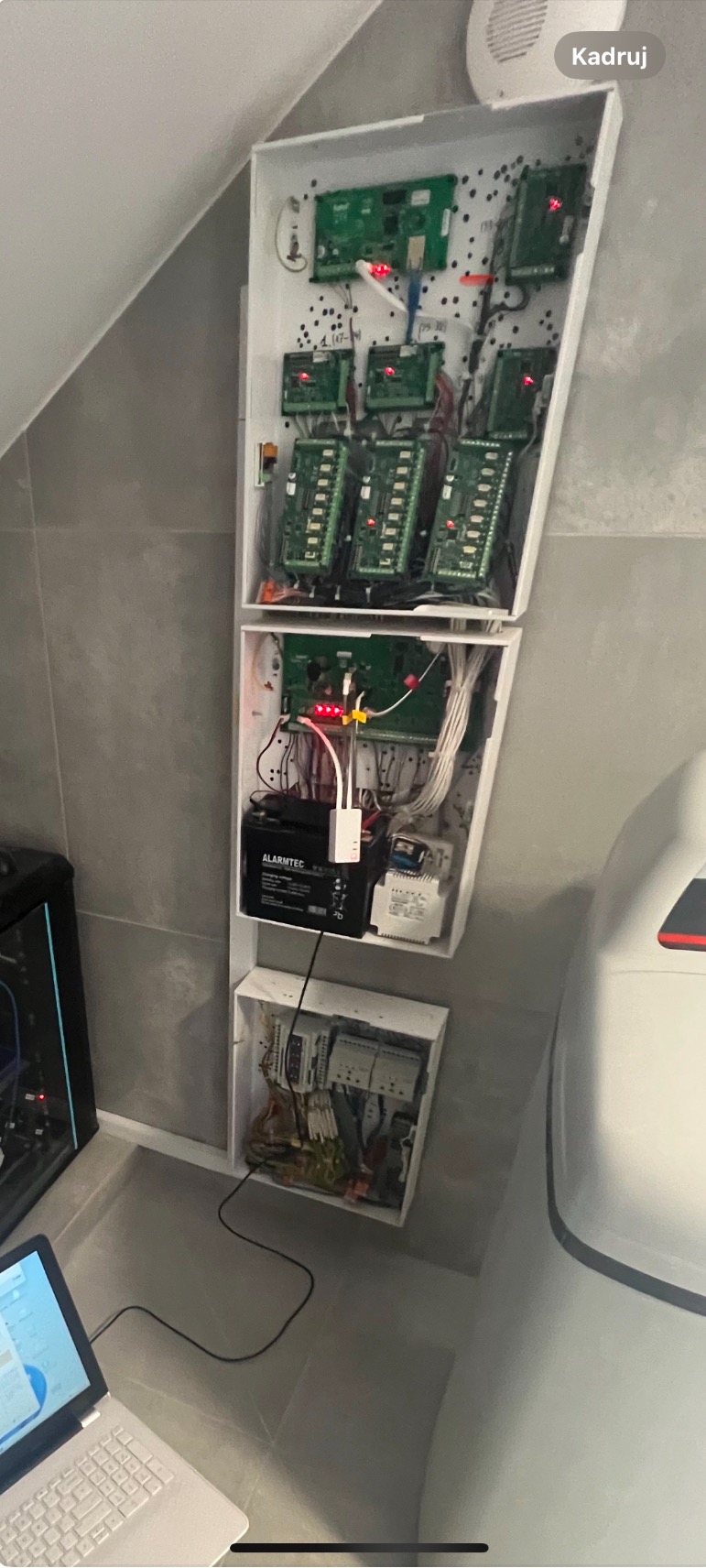 Rozbudowana centrala alarmowa w białej obudowie, z widocznymi płytkami drukowanymi, przewodami i akumulatorem Alarmtec, podłączona do laptopa wyświetlającego dane konfiguracyjne.