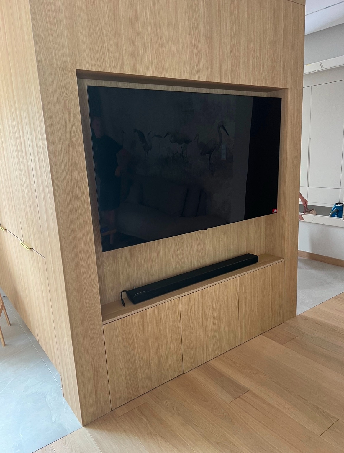 Telewizor wbudowany w drewnianą zabudowę meblową z soundbarem, widoczne drzwi do innego pomieszczenia w tle.