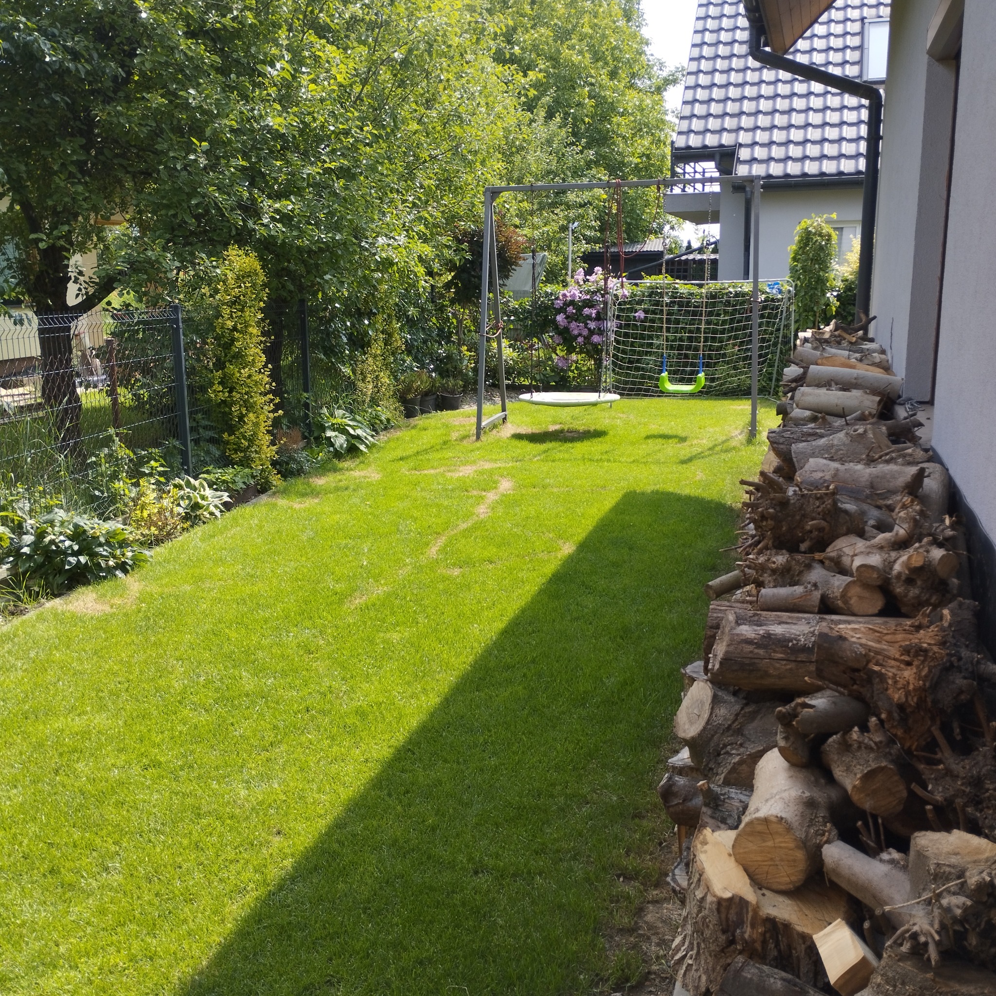 Zadbany trawnik w ogrodzie z huśtawką i stosem drewna przy ścianie domu. Widoczne drzewa, krzewy i ogrodzenie. Cień na trawie.