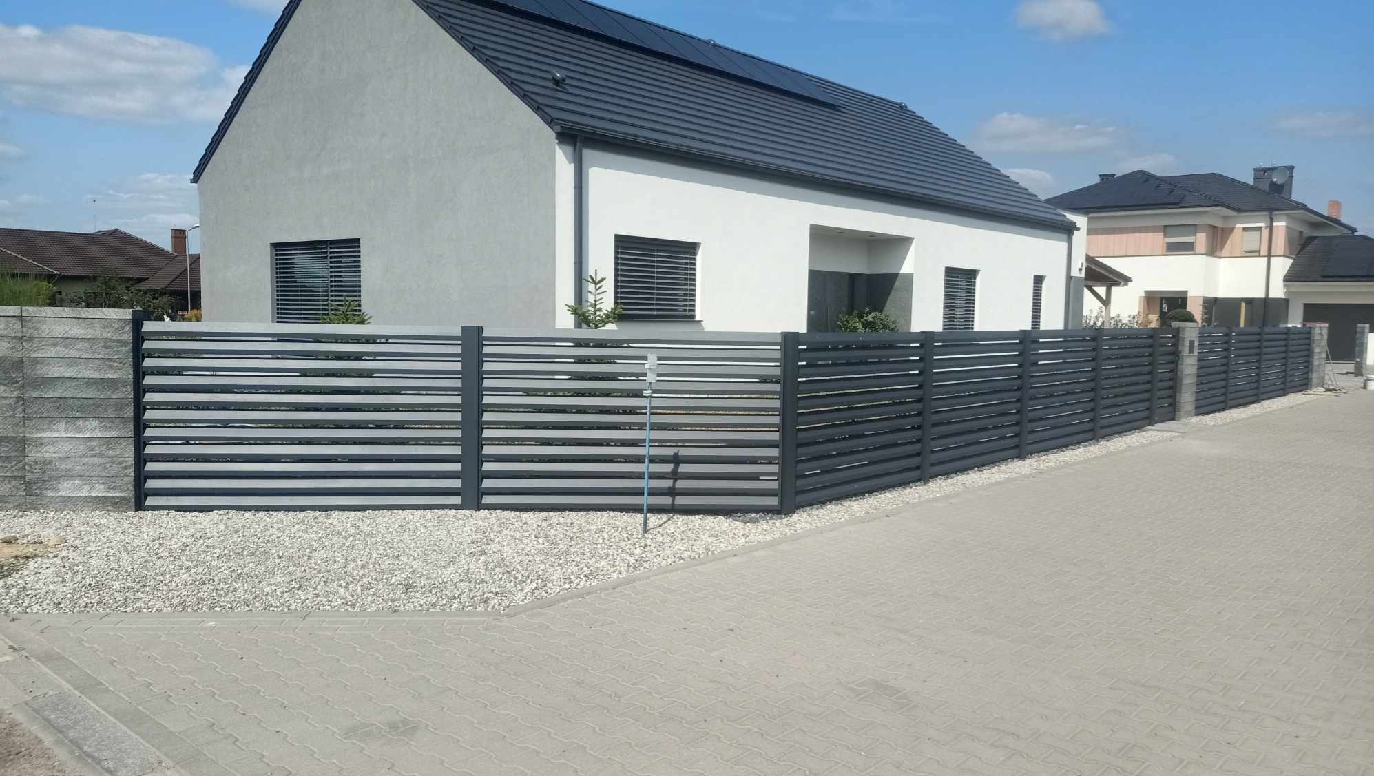 Nowoczesne ogrodzenie panelowe z poziomych listew w kolorze grafitowym, oddzielające posesję z białym domem o minimalistycznej architekturze od chodnika wyłożonego kostką brukową.