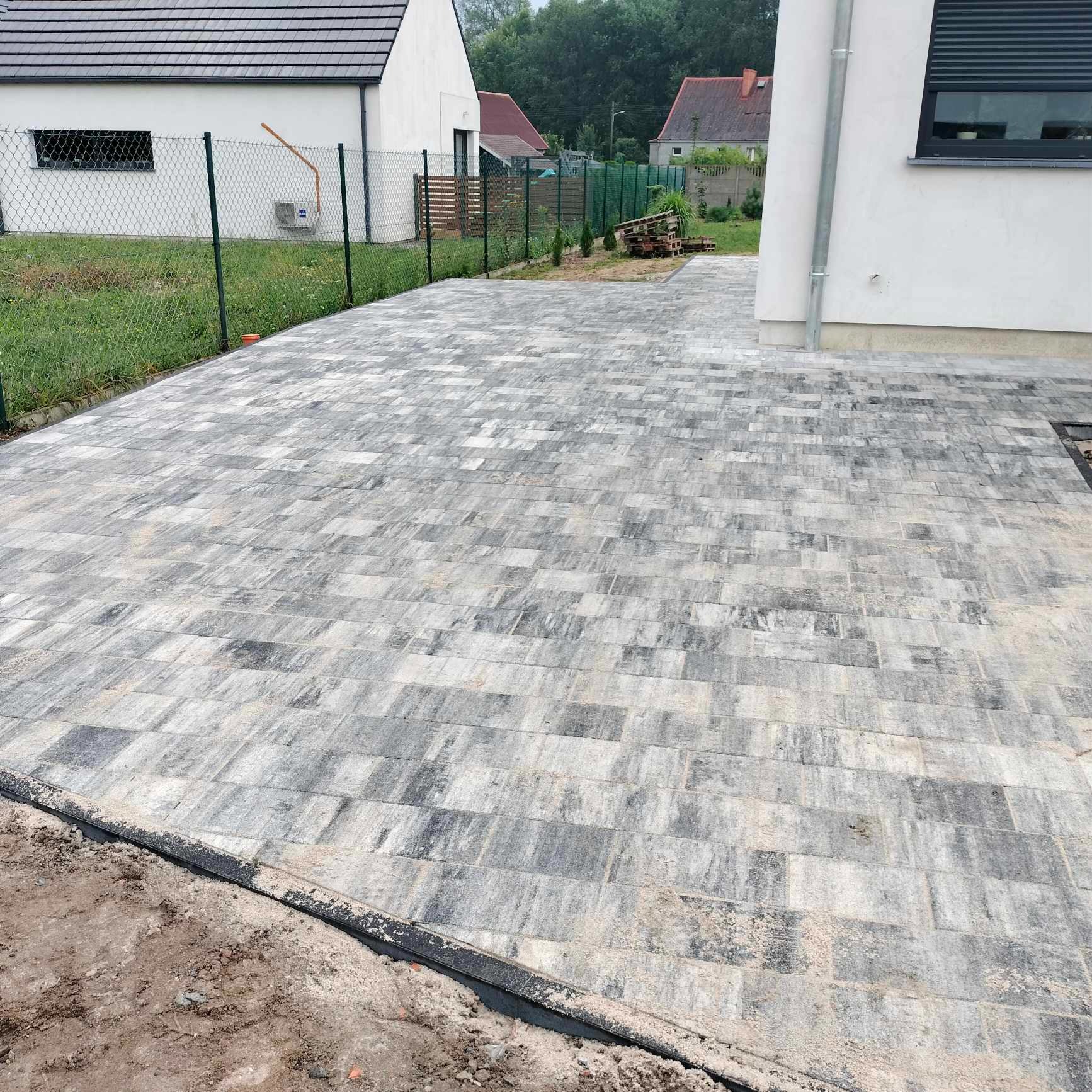 Świeżo ułożona, szara kostka brukowa na podjeździe przed nowoczesnym domem z białymi ścianami i czarnymi roletami, widoczna siatka ogrodzeniowa i trawnik w tle.