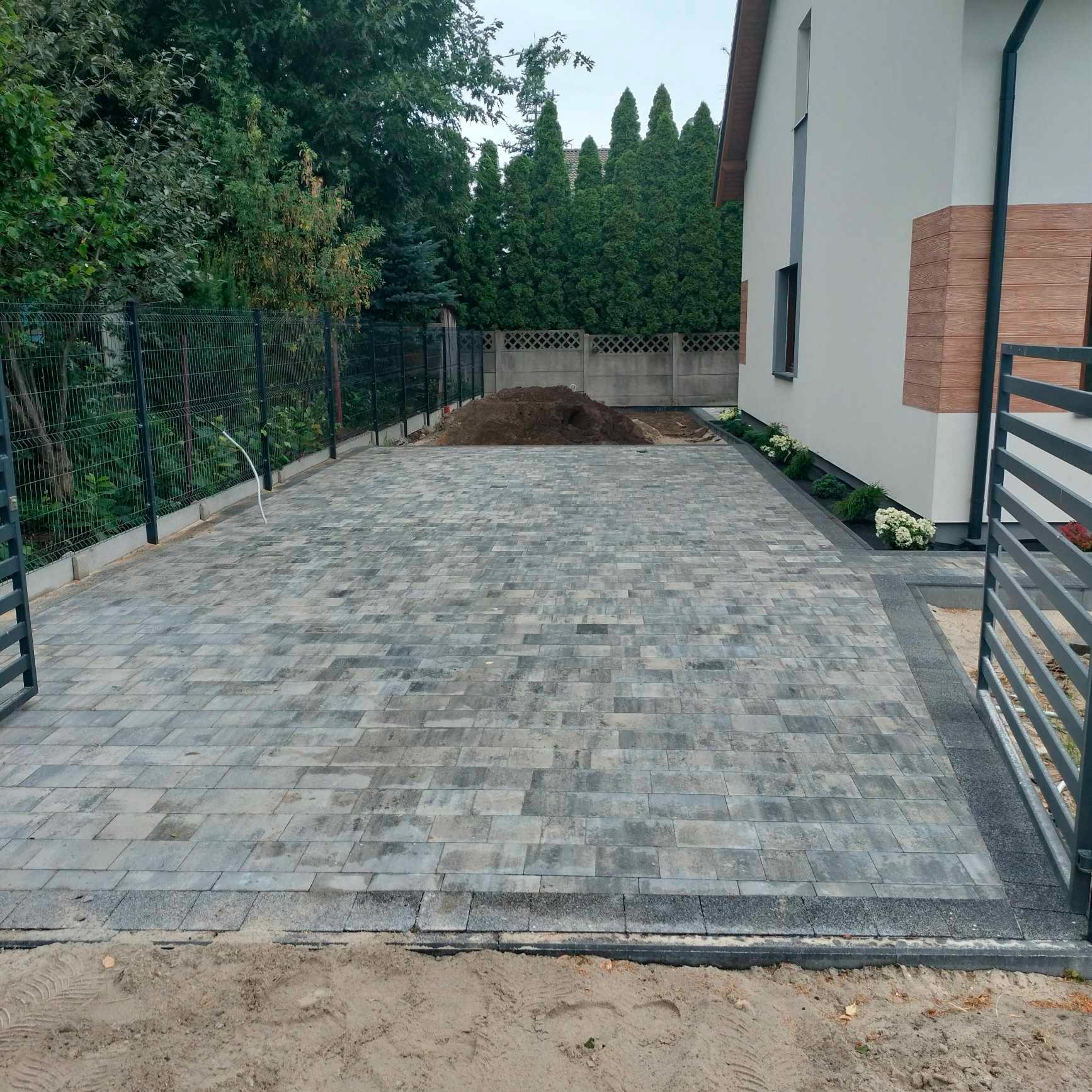 Ułożona kostka brukowa na podjeździe w Kórniku, widok z poziomu gruntu, z fragmentem domu i ogrodzeniem panelowym w tle.