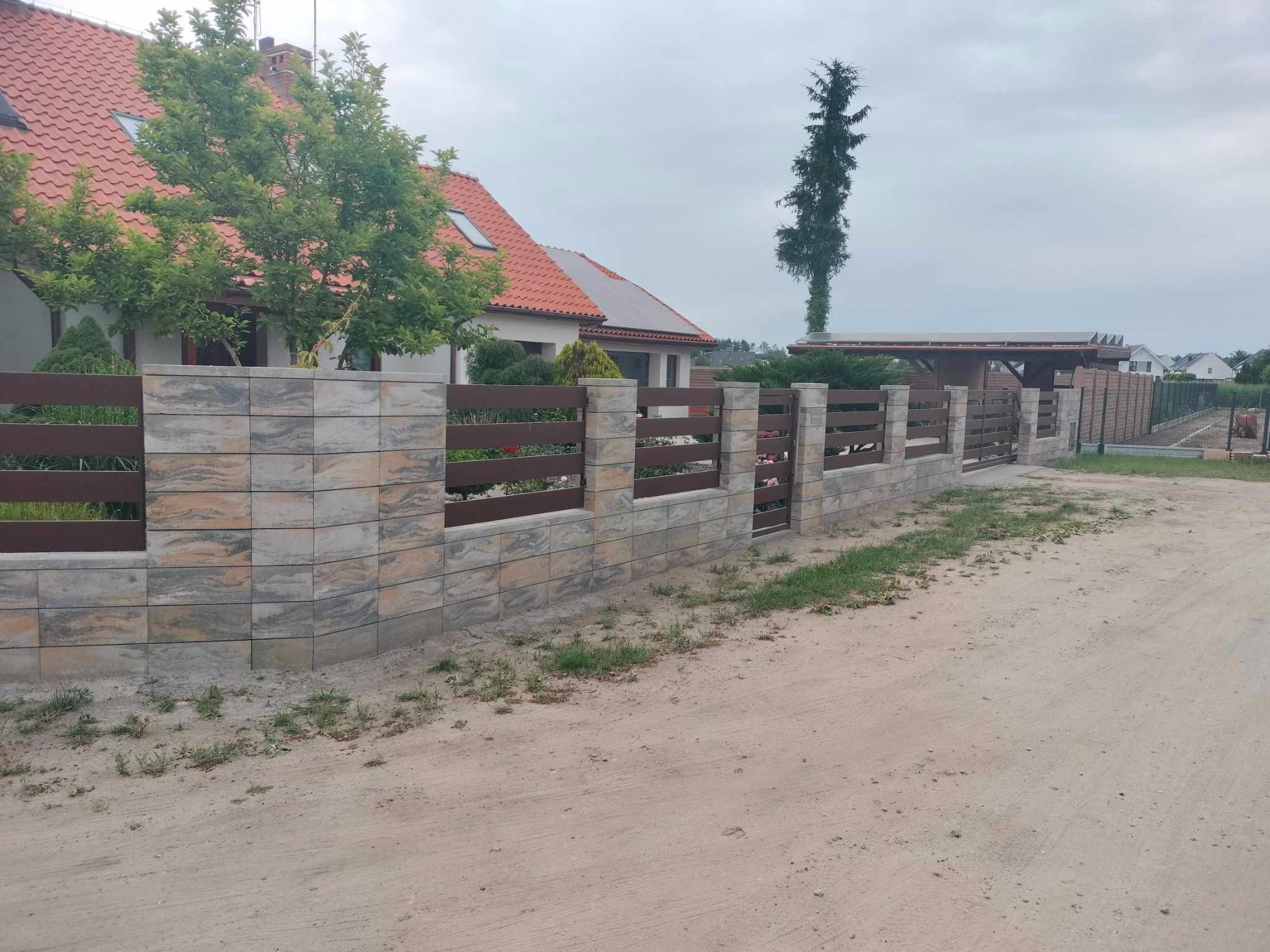 Nowoczesne ogrodzenie panelowe z elementami betonowymi w odcieniach szarości i brązu, z widocznym fragmentem domu jednorodzinnego z czerwoną dachówką w tle.