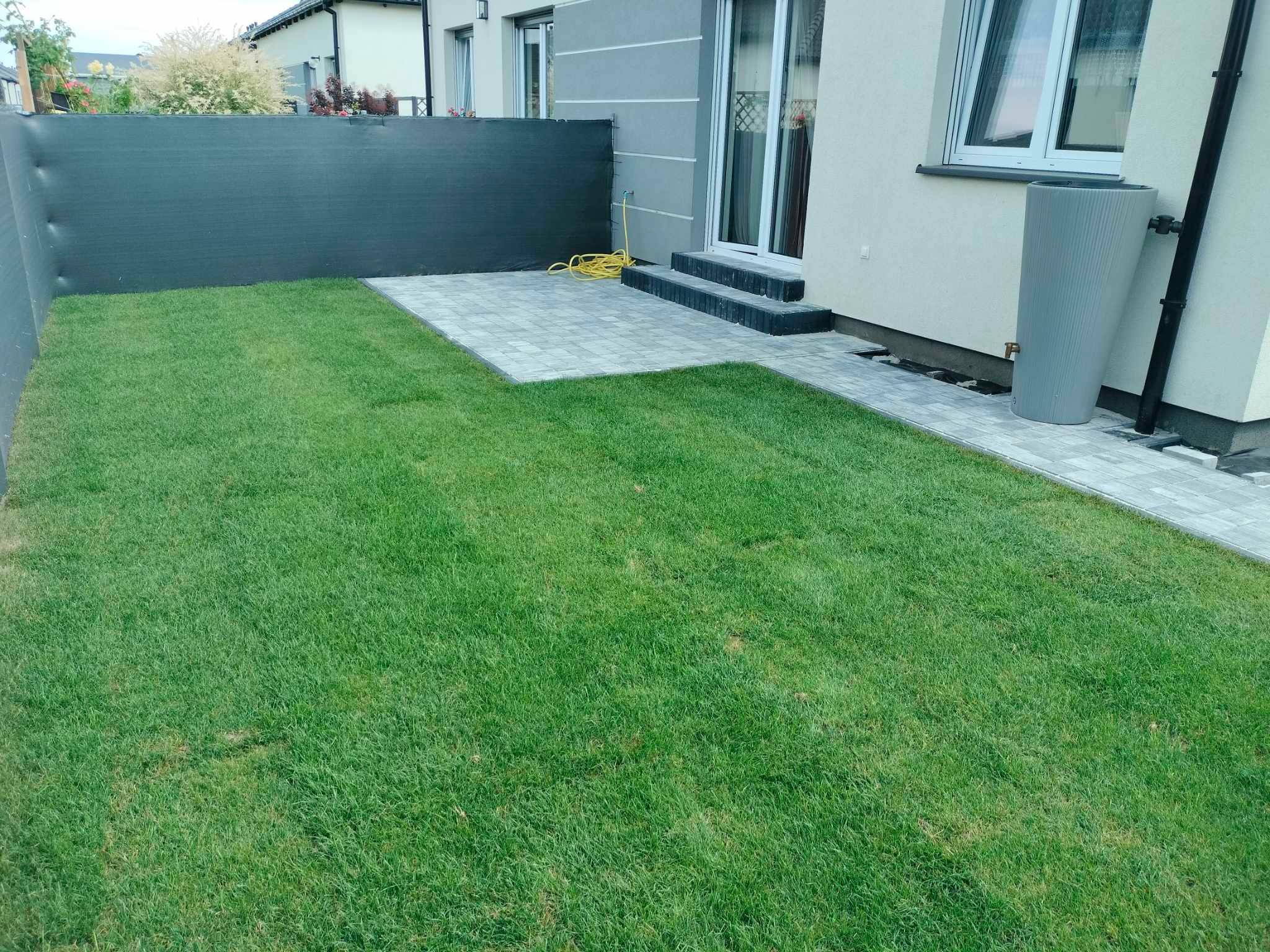 Świeżo ułożona kostka brukowa tworząca ścieżkę i taras przy domu z zielonym trawnikiem, czarną siatką ogrodzeniową i szarym zbiornikiem na deszczówkę.