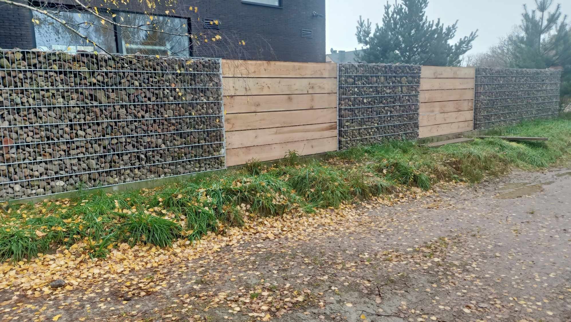 Fragment ogrodzenia z gabionów wypełnionych otoczakami, przeplatanych drewnianymi panelami, widoczne jesienne liście na ziemi.