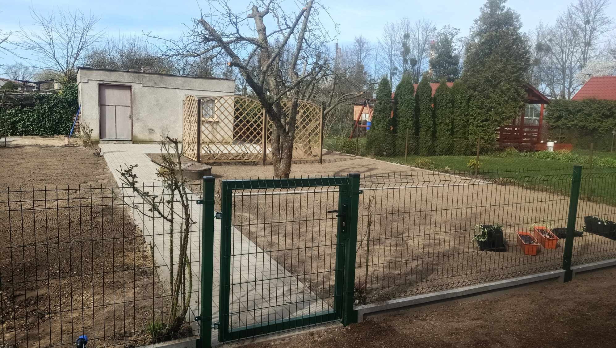Przygotowany teren pod ogród z nową, zieloną bramką panelową i ścieżką z kostki prowadzącą do budynku gospodarczego z kratką wokół drzewa.