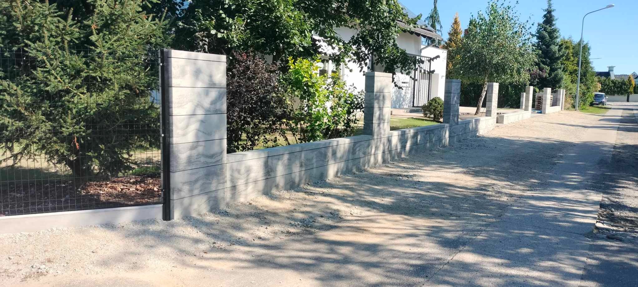 Nowoczesne ogrodzenie panelowe z betonowych elementów, montowane na posesji z widocznymi drzewami i krzewami, w tle biały dom jednorodzinny.