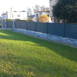 ARTUR MENŻYK - Nowoczesne, poziome ogrodzenie z paneli w kolorze grafitowym, osadzone na niskim murku z kamienia dekoracyjnego, na tle zadbanego trawnika i zabudowy jednorodzinnej.