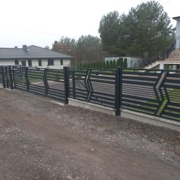 ARTUR MENŻYK - Nowoczesne, poziome ogrodzenie panelowe w kolorze grafitowym z geometrycznym wzorem w kształcie strzałek, oddzielające posesję od żwirowej drogi dojazdowej, z widocznym domem i zielenią w tle.