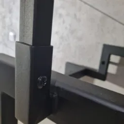 Szczegółowy widok łączenia elementów czarnej, metalowej balustrady z widoczną śrubą mocującą, na tle ściany o fakturze betonu.