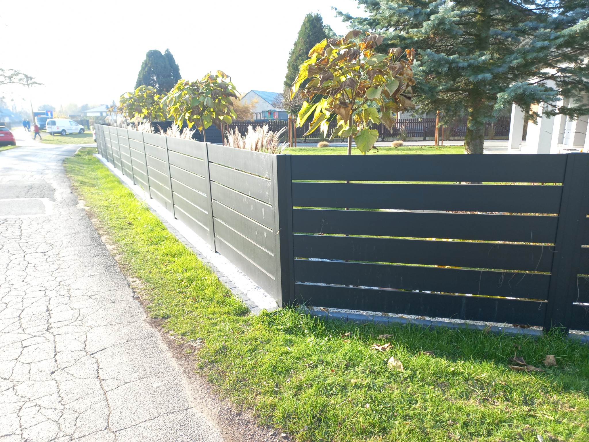 Nowoczesne, poziome przęsła ogrodzeniowe w kolorze grafitowym, oddzielające posesję od chodnika z popękanego asfaltu, z roślinami ozdobnymi posadzonymi wzdłuż ogrodzenia.