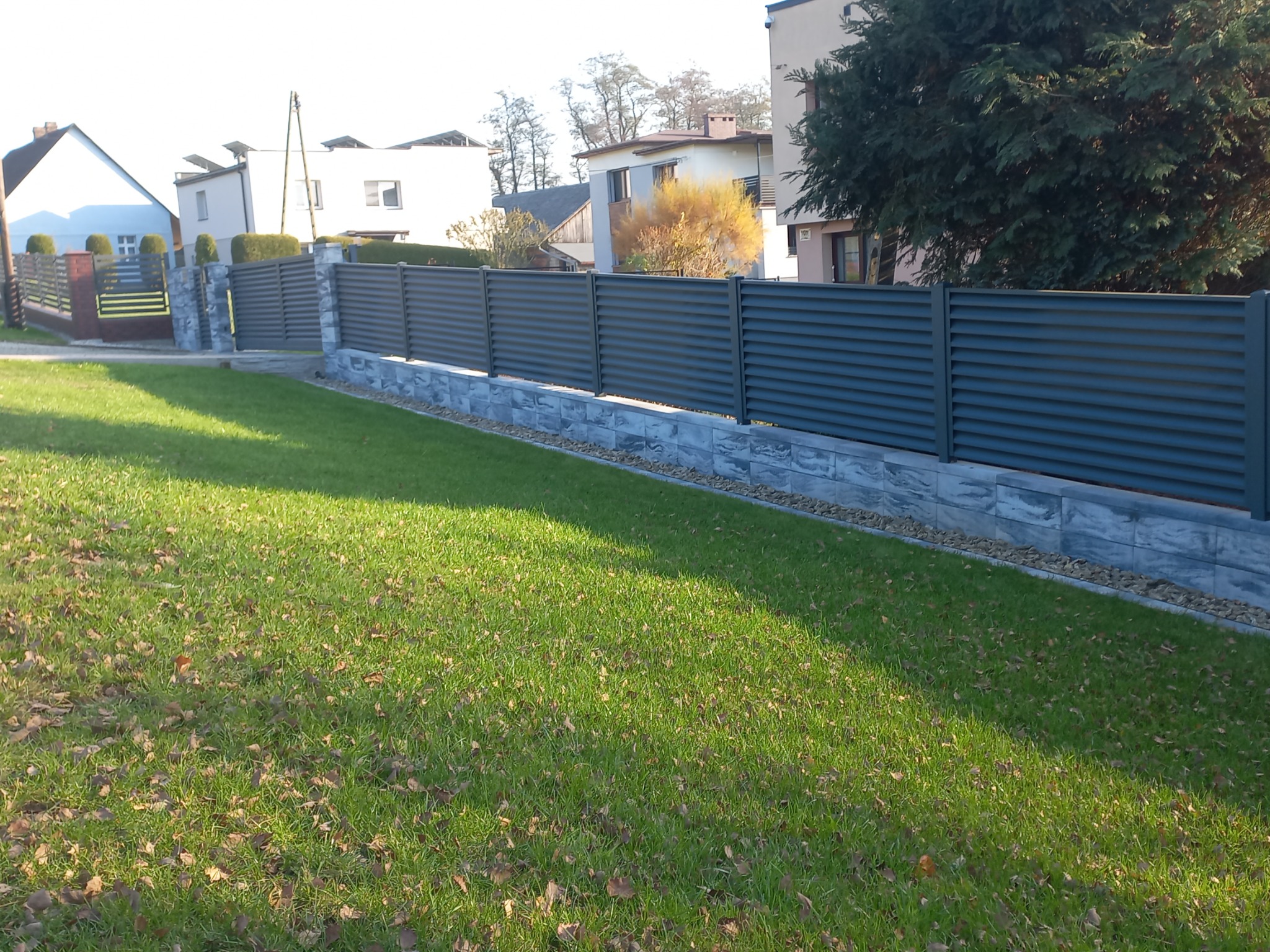 Nowoczesne, poziome ogrodzenie z paneli w kolorze grafitowym, osadzone na niskim murku z kamienia dekoracyjnego, na tle zadbanego trawnika i zabudowy jednorodzinnej.
