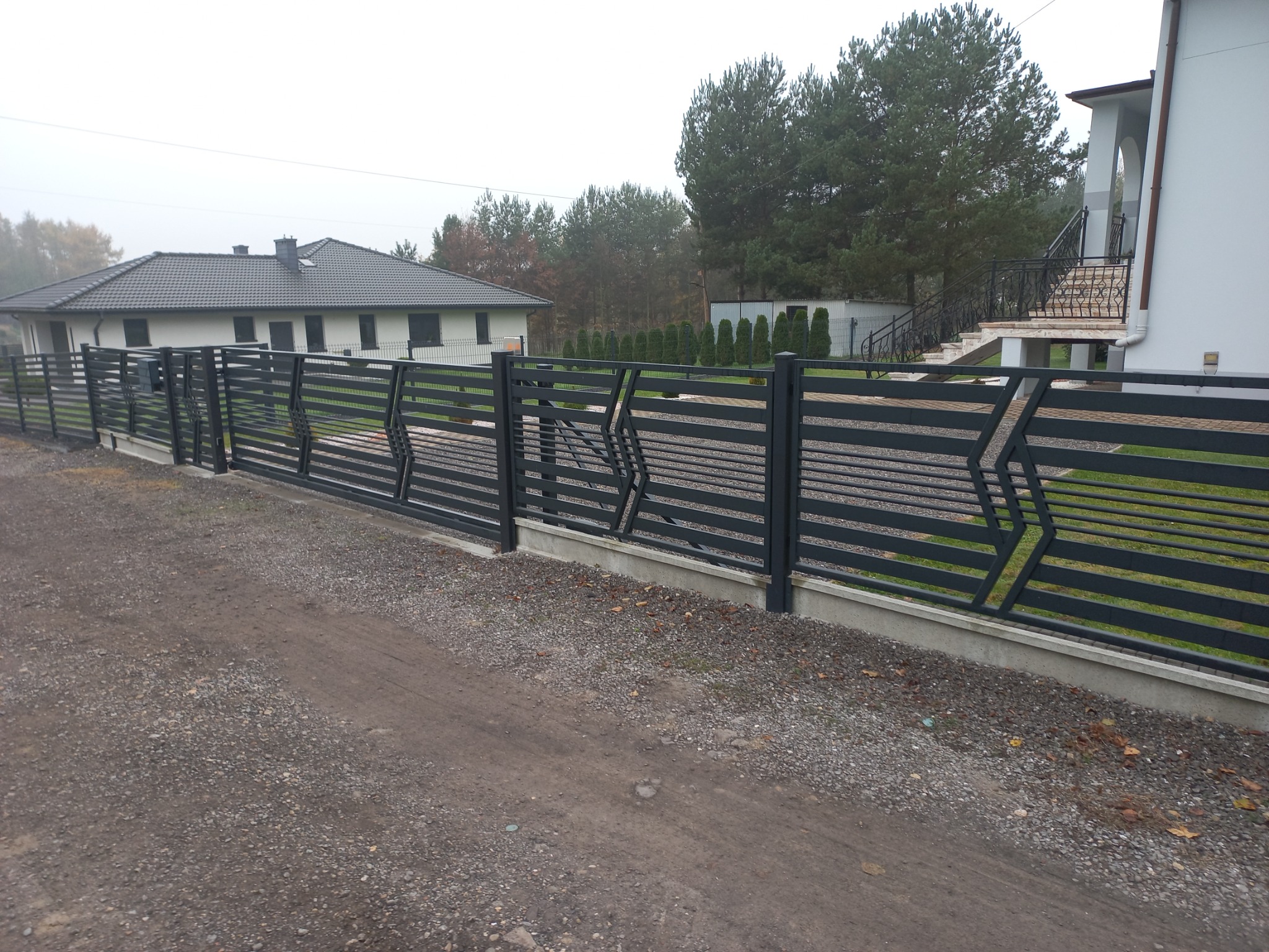 Nowoczesne, poziome ogrodzenie panelowe w kolorze grafitowym z geometrycznym wzorem w kształcie strzałek, oddzielające posesję od żwirowej drogi dojazdowej, z widocznym domem i zielenią w tle.