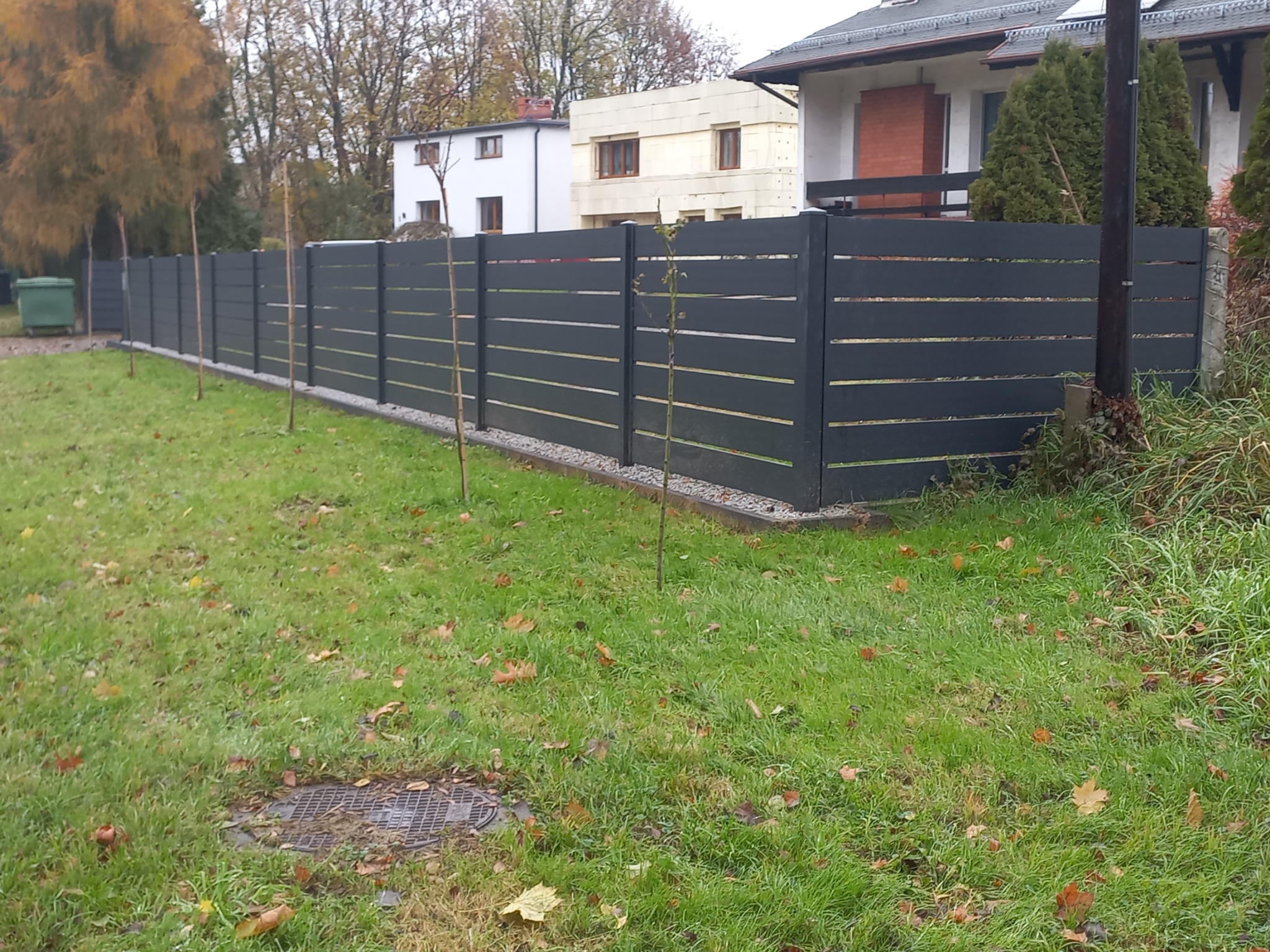 Nowoczesne, poziome ogrodzenie z szarych paneli oddzielające posesję z trawnikiem od sąsiednich domów, z młodymi drzewkami posadzonymi wzdłuż linii ogrodzenia.