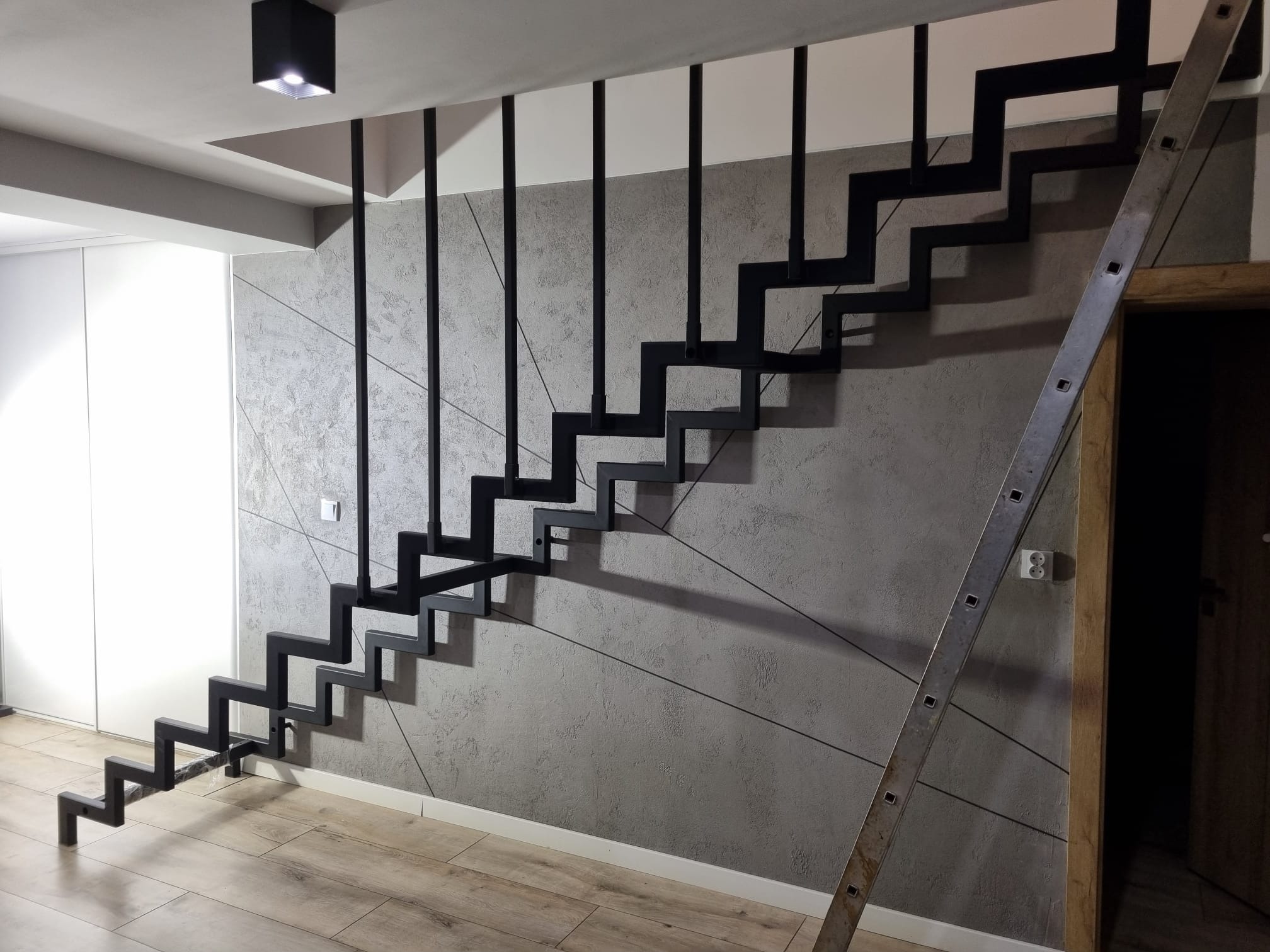 Nowoczesne, minimalistyczne schody metalowe o konstrukcji wspornikowej, pomalowane na czarno, zamontowane przy ścianie z dekoracyjnym tynkiem. W tle widoczna drabina oparta o ścianę.