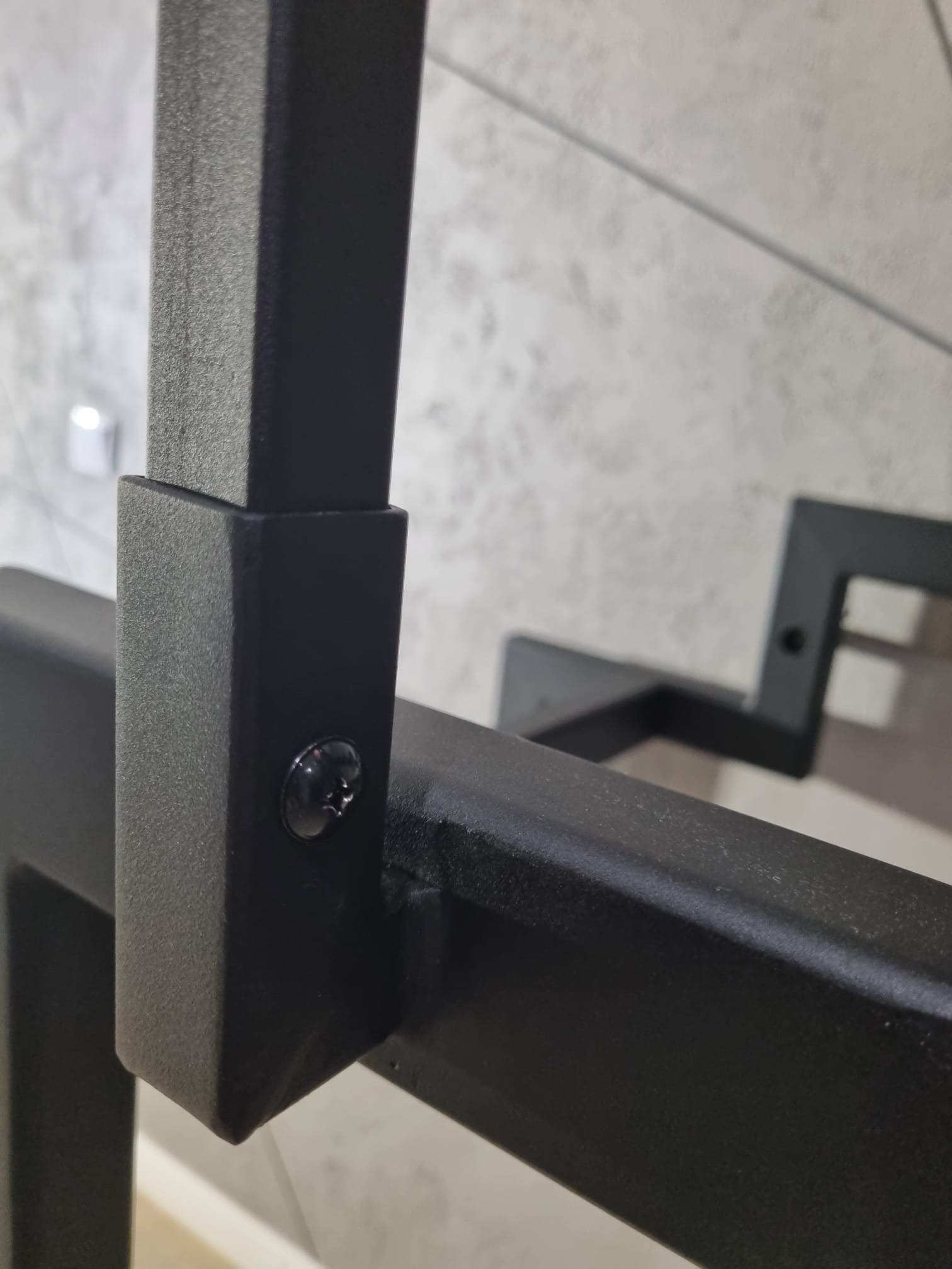 Szczegółowy widok łączenia elementów czarnej, metalowej balustrady z widoczną śrubą mocującą, na tle ściany o fakturze betonu.