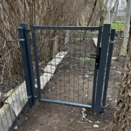 Otwarta, grafitowa furtka z siatki z zamkiem, zamontowana między drzewami i betonowym obrzeżem w ogrodzie.