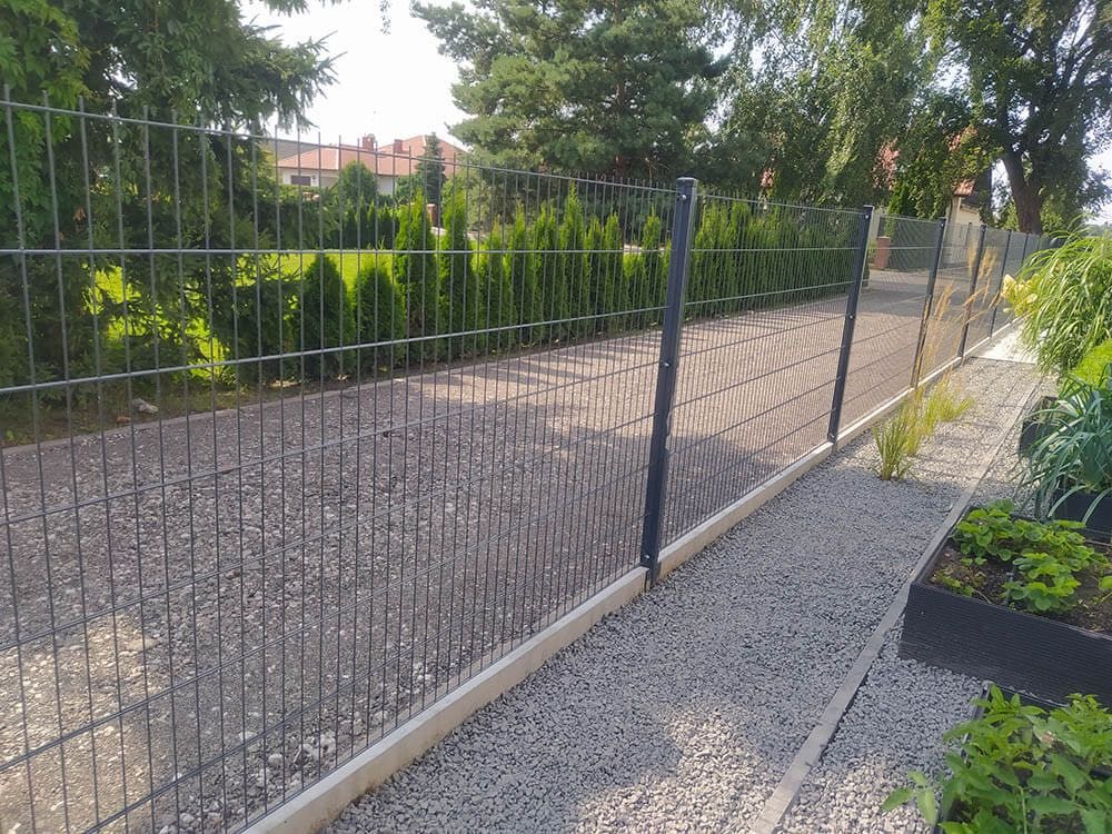 Szary panelowy system ogrodzeniowy z podmurówką betonową, widok z boku na żwirową ścieżkę i rabaty roślinne, w tle zielone tuje i dom.