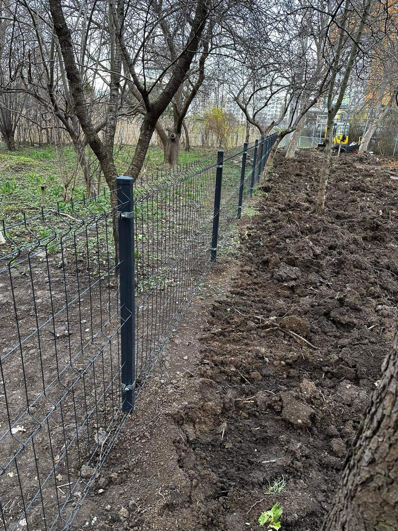 Nowo zainstalowane, czarne, metalowe ogrodzenie z siatki panelowej na granicy działki, z widoczną świeżo przekopaną ziemią i sprzętem budowlanym w tle.