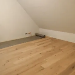 Jasne panele podłogowe ułożone w pokoju na poddaszu, widoczna rolka podkładu pod panele częściowo rozwinięta przy ścianie.