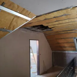 Poddasze w trakcie ocieplania wełną mineralną, widoczne metalowe profile konstrukcyjne, okno dachowe i przygotowane ściany do wykończenia.