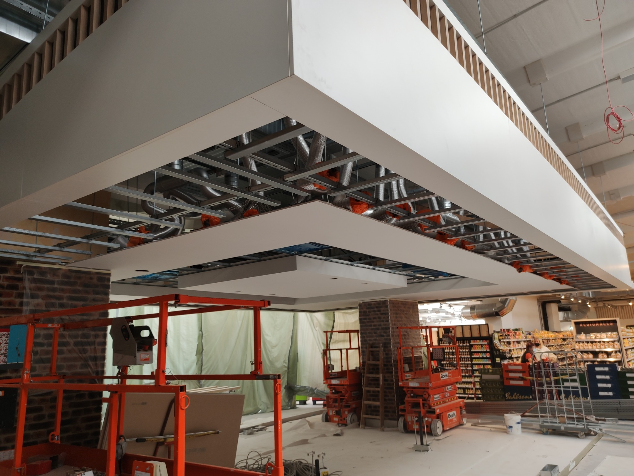 Montaż podwieszanego sufitu z widoczną konstrukcją metalową i instalacjami w przestrzeni handlowej, z użyciem podnośników koszowych.