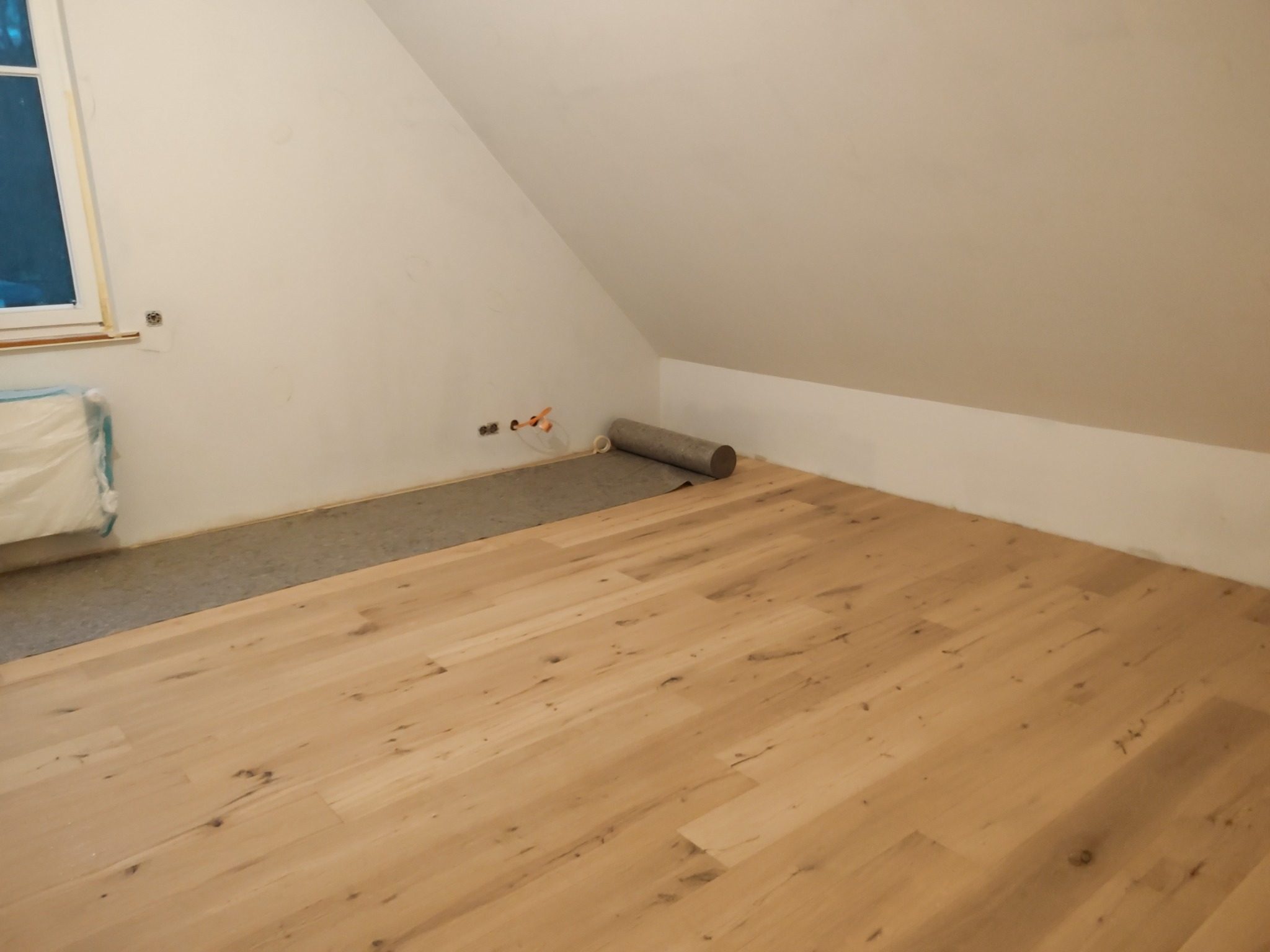 Jasne panele podłogowe ułożone w pokoju na poddaszu, widoczna rolka podkładu pod panele częściowo rozwinięta przy ścianie.