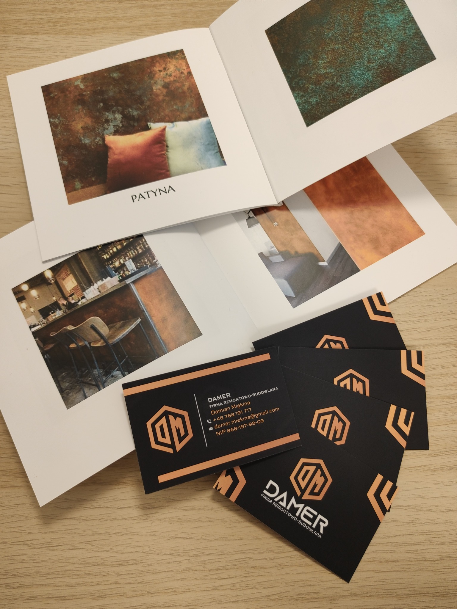 Katalog z inspiracjami wykończenia ścian z efektem patyny i wizytówki firmy remontowej DAMER na drewnianym biurku.