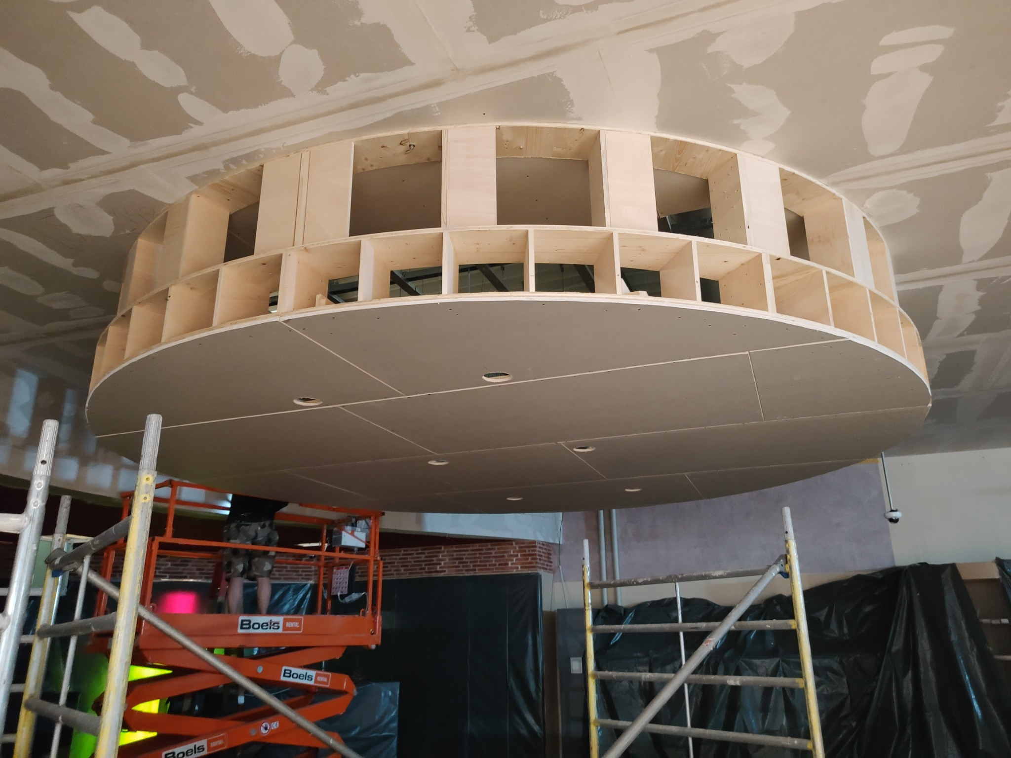 Niedokończona okrągła konstrukcja z płyt kartonowo-gipsowych i drewnianych belek, zamontowana pod sufitem, widoczna z dołu z podnośnika i drabiny w tle.