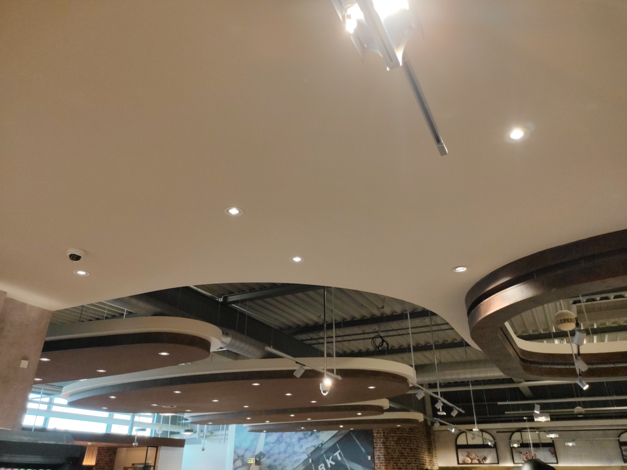 Widok na sufit w dużym pomieszczeniu z dekoracyjnymi elementami podwieszanymi, oświetleniem punktowym i widocznymi instalacjami technicznymi.