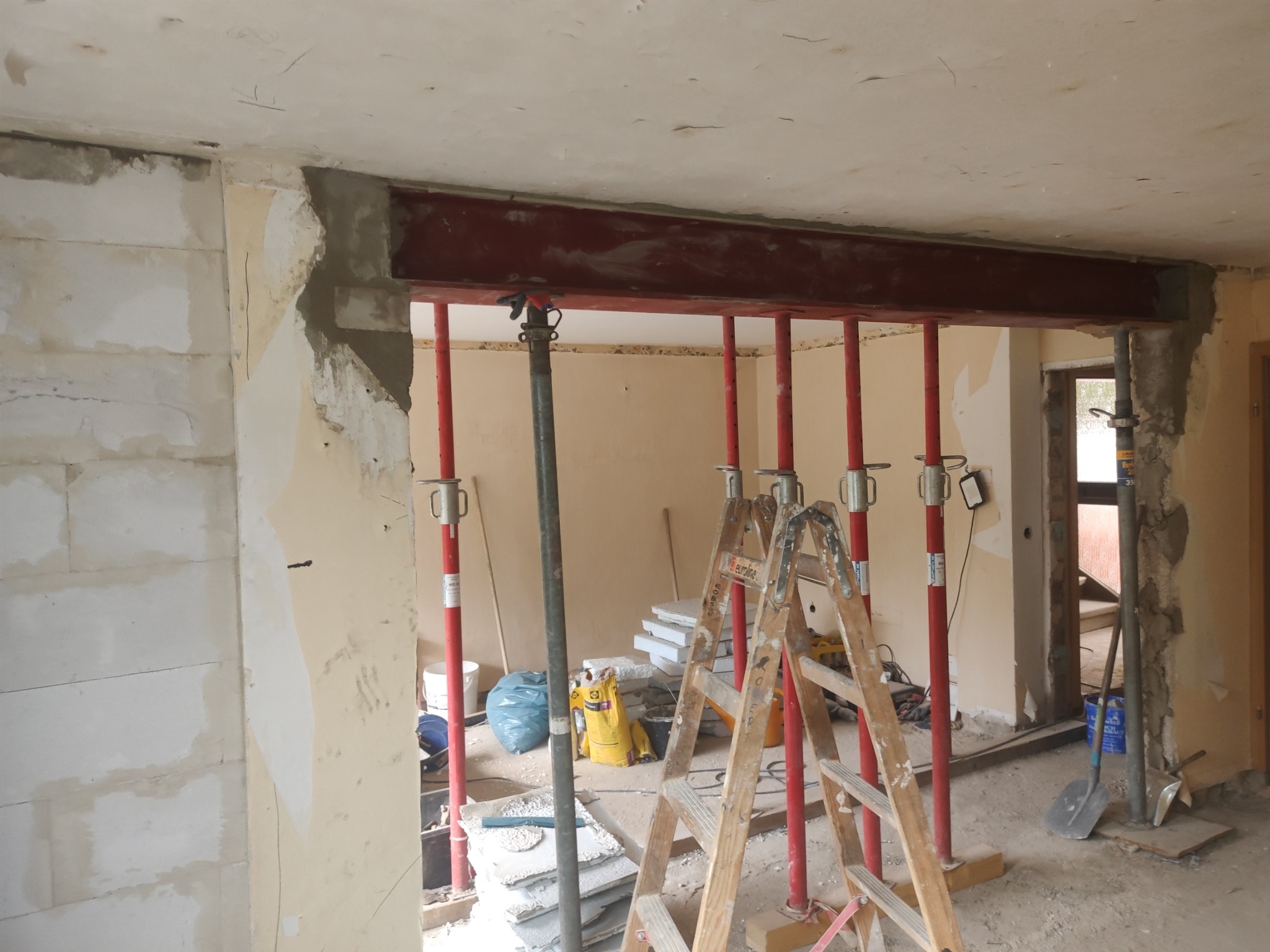 Wzmocnienie ściany nośnej za pomocą stalowej belki podpartej czerwonymi stemplami, w tle drabina i materiały budowlane.