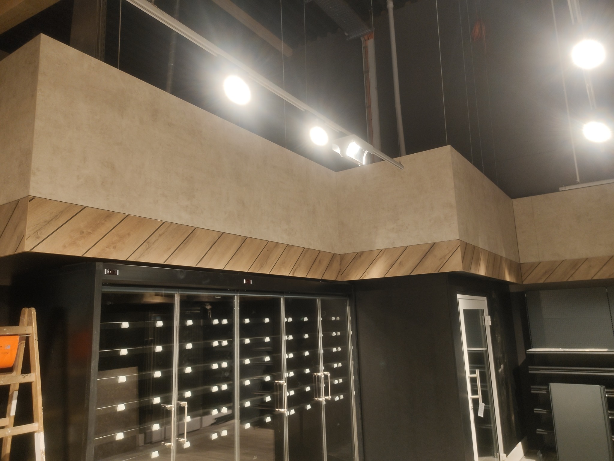 Wykończenie wnętrza sklepu z beżowymi ścianami, drewnianą listwą dekoracyjną i przeszklonymi drzwiami, oświetlone punktowymi lampami.