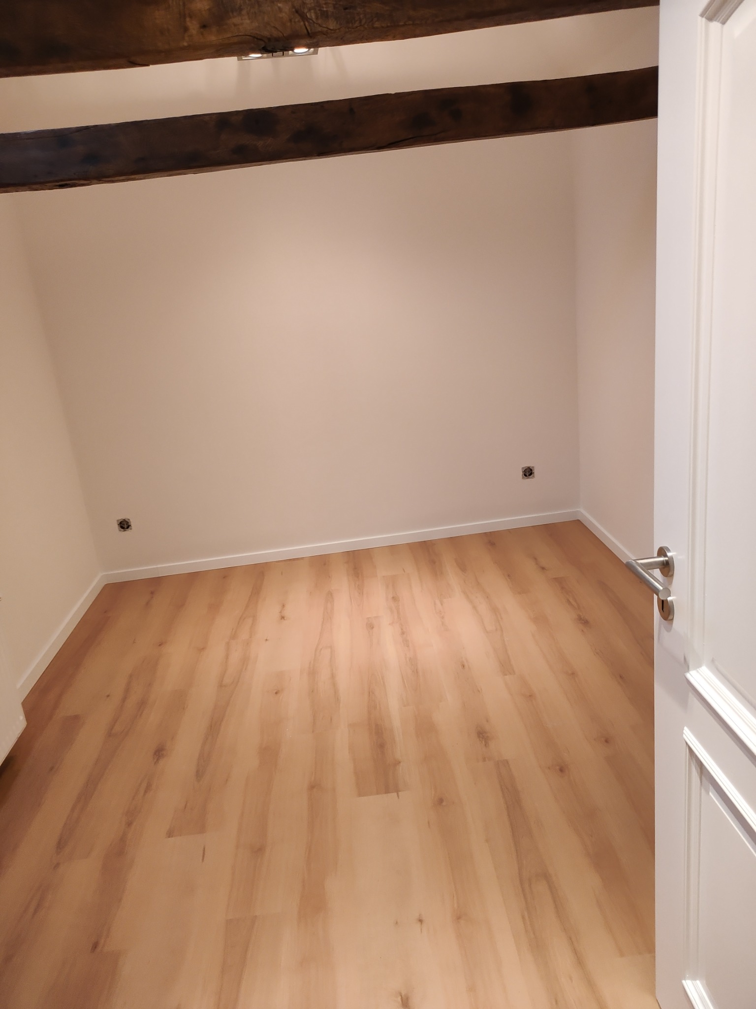 Wnętrze pokoju z nową, jasną podłogą z paneli, widoczne drewniane belki stropowe i białe ściany z gniazdkami elektrycznymi, fragment białych drzwi po prawej stronie.