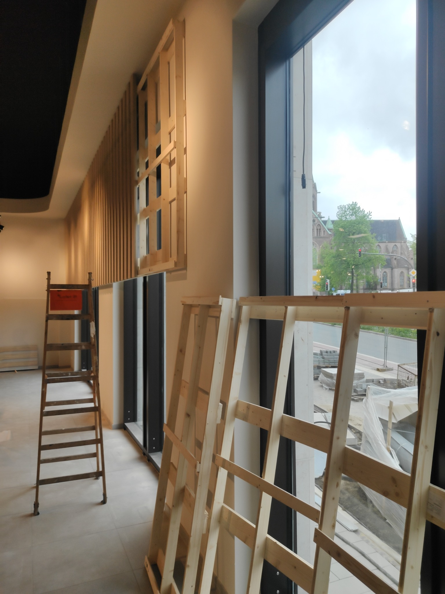 Wnętrze w trakcie prac: drewniana drabina oparta o ścianę, drewniane ramy okienne ustawione w pomieszczeniu, widok na budynek kościoła przez okno.