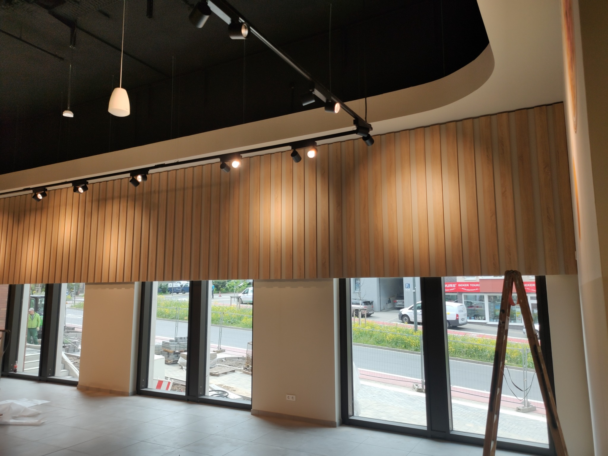 Wnętrze lokalu w trakcie prac wykończeniowych: drewniana okładzina ścienna, system oświetlenia szynowego, duże okna z widokiem na ulicę, drabina oparta o ścianę, posadzka z płytek.