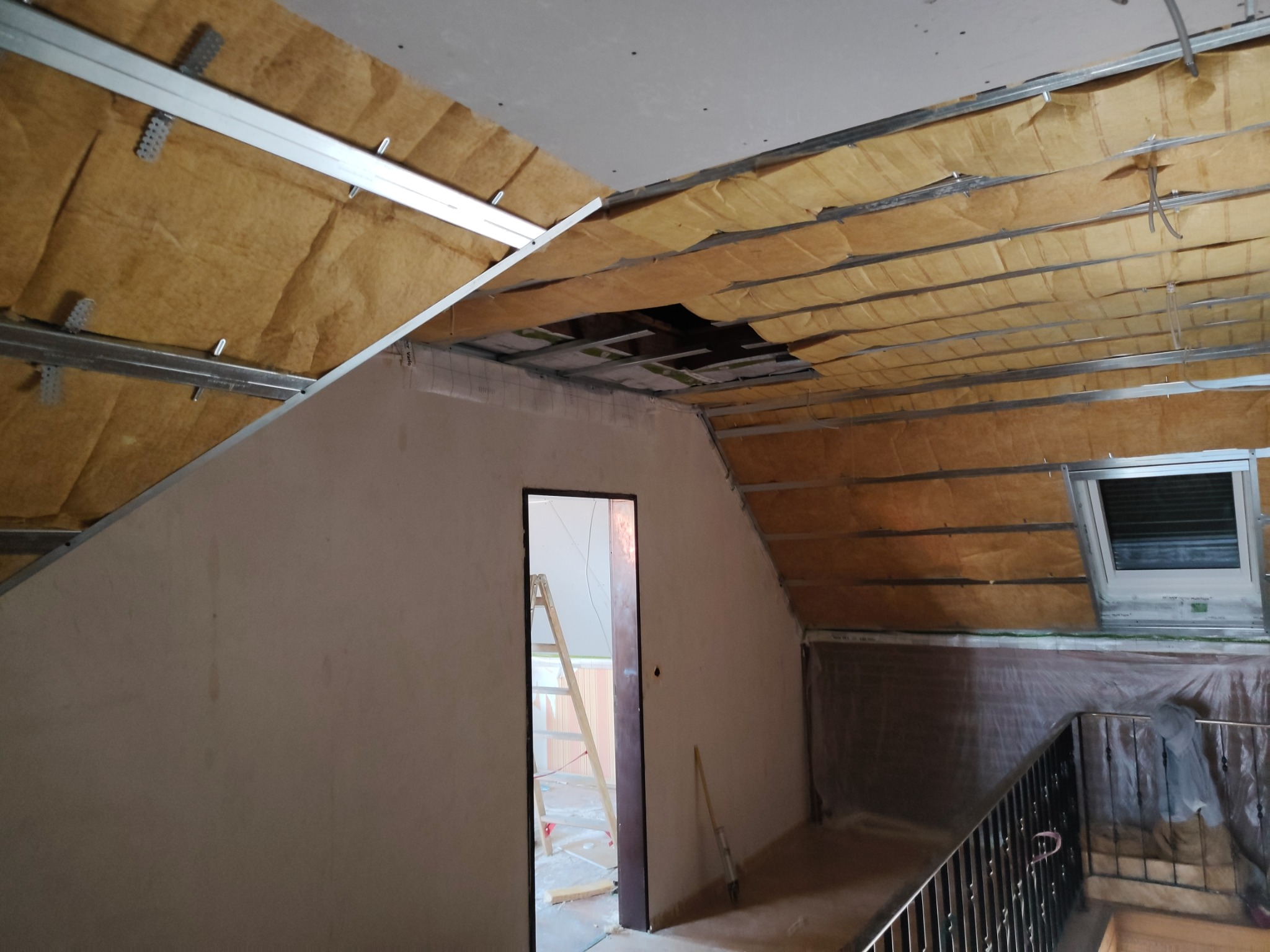 Poddasze w trakcie ocieplania wełną mineralną, widoczne metalowe profile konstrukcyjne, okno dachowe i przygotowane ściany do wykończenia.