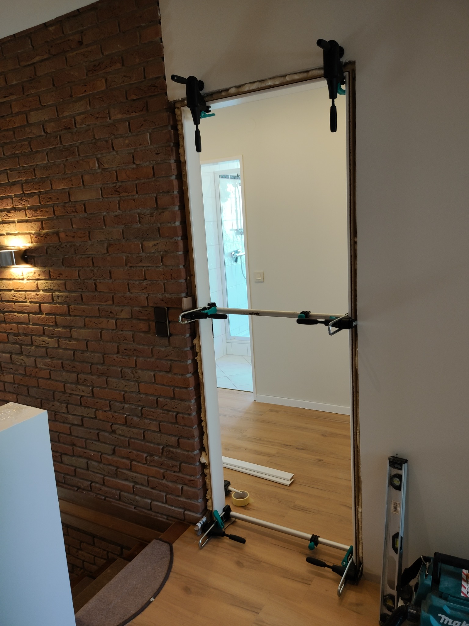 Montaż ościeżnicy drzwiowej z użyciem rozpórki teleskopowej, widoczna ściana z cegły i poziomica