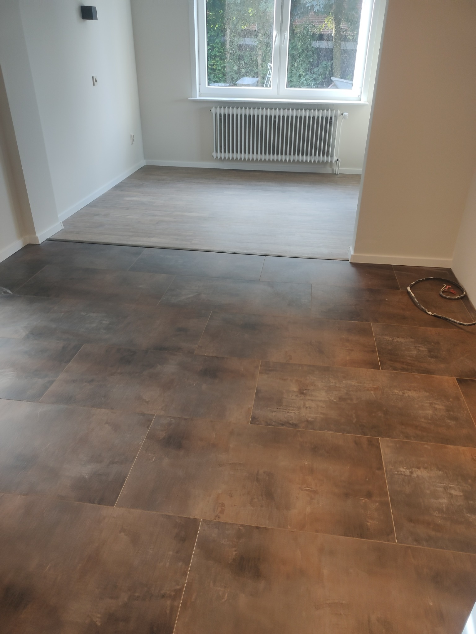 Ułożone brązowe płytki podłogowe w nowym mieszkaniu w Rzezawie, widoczny fragment drugiego pomieszczenia z szarą podłogą i grzejnikiem pod oknem.