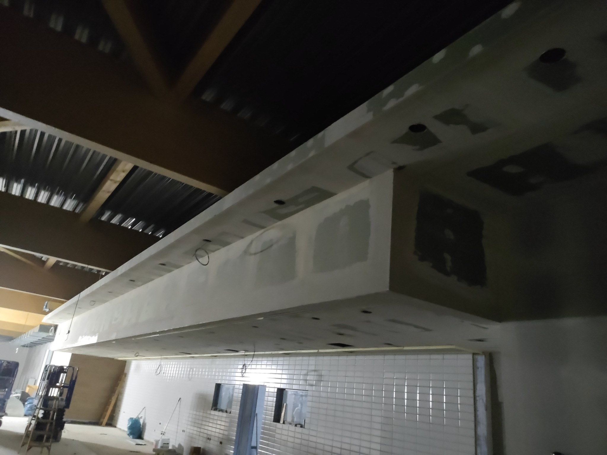 Sufit podwieszany z płyt kartonowo-gipsowych w trakcie szpachlowania, widoczne otwory na oświetlenie, ściany wyłożone białymi płytkami, surowy stan wykończenia wnętrza.